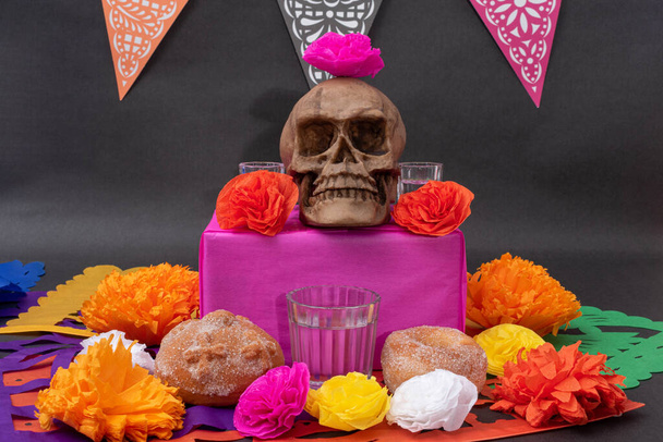Mexikói Day of the dead altar with traditional bread, colorful flowers, banner, bread, mezcal, a skull and cut paper on black background - Fotó, kép