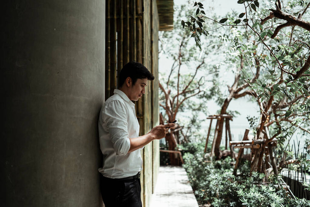 Maduro asiático hombre de negocios en blanco formal camisa apoyada en un palo jugando con el teléfono inteligente en el jardín
. - Foto, Imagen