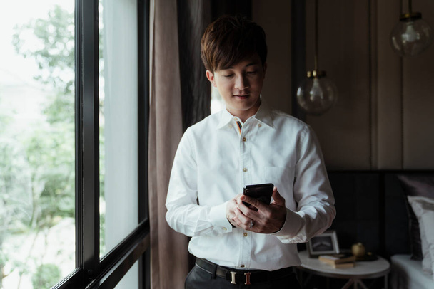 Азиатский мужчина в белой рубашке с помощью смартфона в комнате рядом с окном
. - Фото, изображение