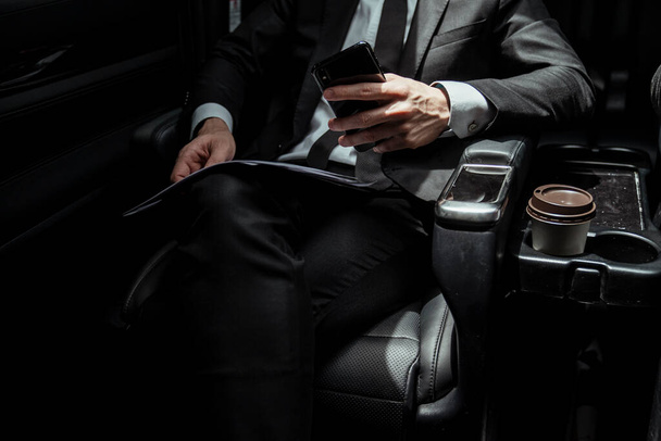 会議への途中でスマートフォンを保持車の中で黒のスーツのシットン上のビジネスマン. - 写真・画像