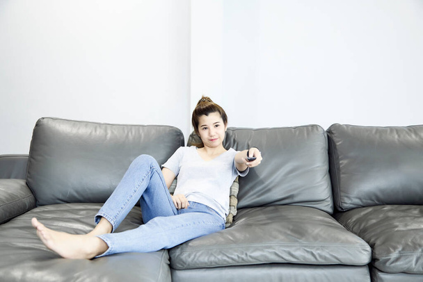 Молодая женщина отдыхает дома, смотрит телевизор и меняет канал с помощью пульта дистанционного управления
 - Фото, изображение