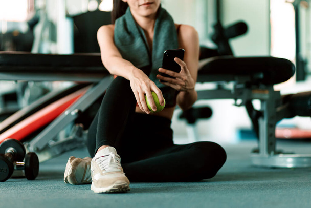 A girl hold green apple using smartphone in fitness gym. Woman exercise in gym fitness breaking and relax. - Photo, Image