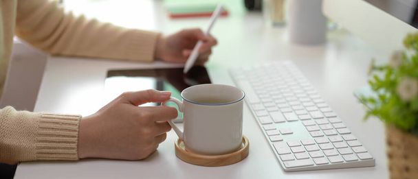 Vue latérale d'une travailleuse travaillant sur une tablette et tenant une tasse de café sur un bureau blanc dans une pièce de bureau  - Photo, image