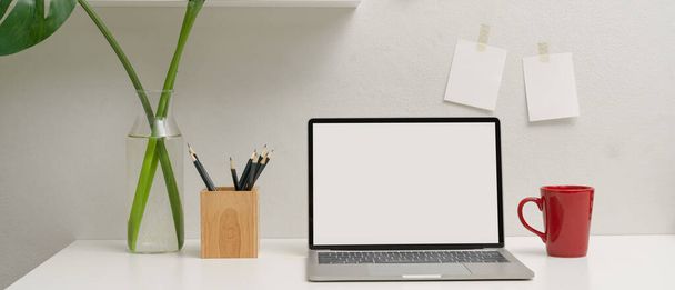 Vista de cerca del espacio de trabajo simple con el ordenador portátil de la maqueta, taza, lápices y jarrón de la planta en la mesa blanca
 - Foto, Imagen