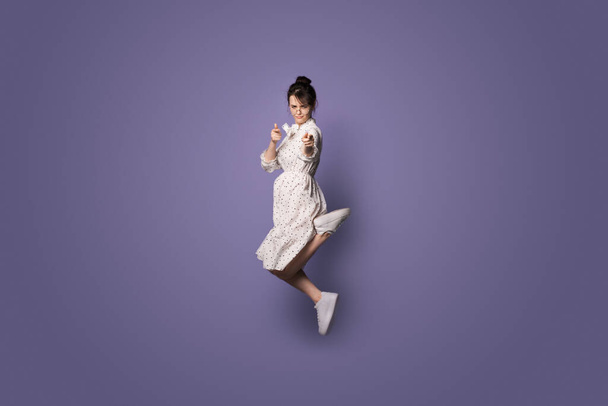 Mulher caucasiana grave pulando em uma parede de estúdio violeta com espaço livre e apontando para a câmera
 - Foto, Imagem