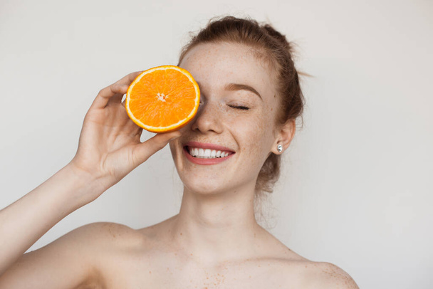 Toothily smiling caucasian lady with freckles and red hair is covering her eye with a sliced orange and posing with naked shoulders on white studio wall - Photo, Image