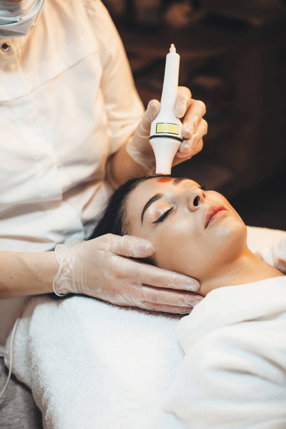 Foto vista lateral de uma mulher caucasiana deitada no salão de spa com um procedimento facial anti envelhecimento com aparato moderno
 - Foto, Imagem