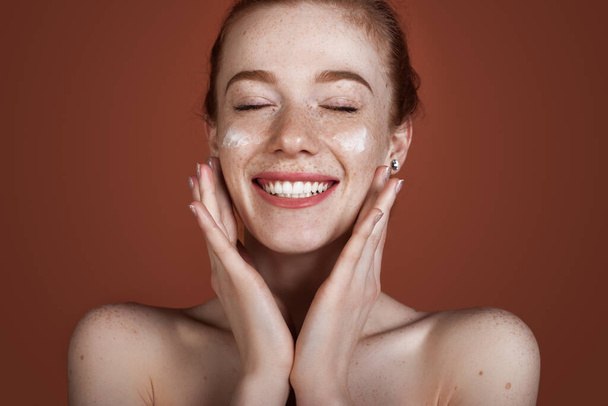 Smiling caucasian ginger lady with freckles applying anti aging cream on face and smile posing with naked shoulders on red studio wall - Foto, afbeelding