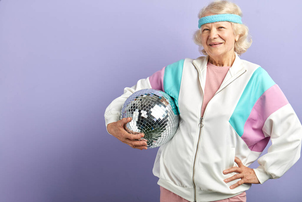 Positive elderly lady holds sparkling disco ball, dressed smiling at camera. - Photo, Image