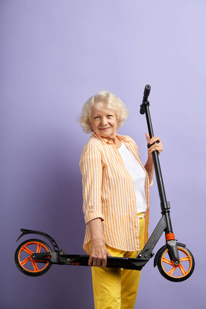 Fun activity. Joyful positive mature woman holding scooter while posing indoors. - Foto, afbeelding