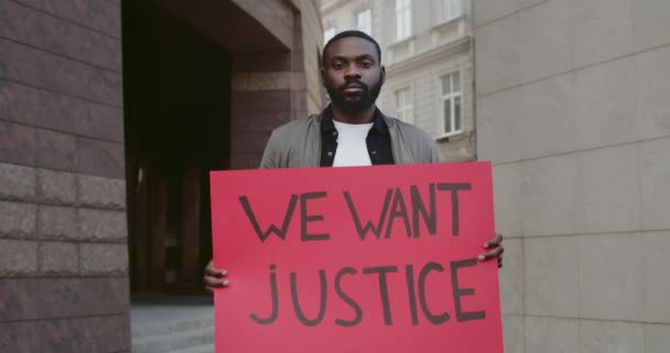 Portrait of guy activist holding banner with we want justice writing on it. Afro american bearded man supporting human rights movement while standing at empty city street. - Πλάνα, βίντεο