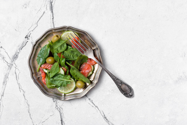Top view of vintage silver plate with red fish, spinach leaves, green olives, lime and cucumber on white marble background with copy space - Fotó, kép