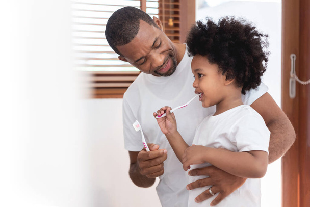 Happy African American Family, Dad teaching his son in white shirts brushing teeth in bathroom together. Father and kid boy in curly hair enjoying with dental hygiene at home in morning - Fotografie, Obrázek