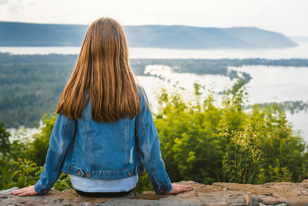 Ένα μικρό κορίτσι ένα παιδί με τζιν μπουφάν κάθεται στην άκρη ενός γκρεμού και κοιτάζει την καταπληκτική φυσική εικόνα με τα βουνά και ένα ποτάμι - Φωτογραφία, εικόνα