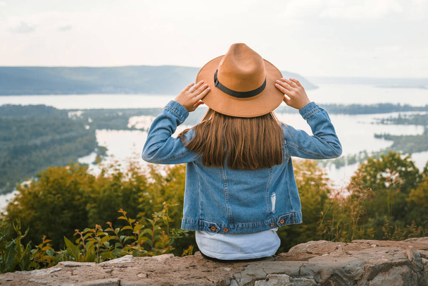 Malá holčička dítě v džínové bundě sedí na okraji útesu drží klobouk v rukou a dívá se na úžasné přírodní obraz s horami a řekou - Fotografie, Obrázek