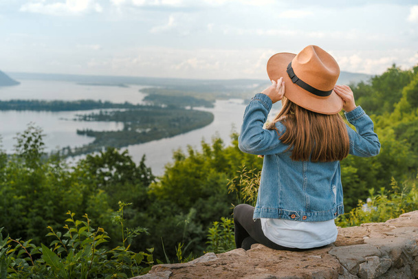 Ένα κοριτσάκι με τζιν μπουφάν κάθεται στην άκρη ενός γκρεμού κρατώντας το καπέλο της στα χέρια της και κοιτάζει την καταπληκτική φυσική εικόνα με τα βουνά και το ποτάμι. - Φωτογραφία, εικόνα