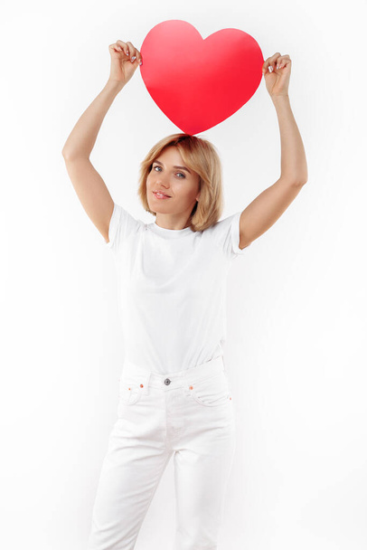 Charming young blonde woman in casual white outfit holding paper heart on the head over white background. - Foto, Imagem