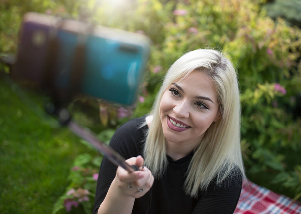 Tini lány szőke haj csinál szelfi mobiltelefon bottal piknik a parkban - Fotó, kép