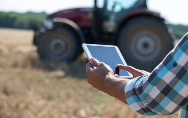 Close up da mão do agricultor segurando tablet na frente do trator que trabalha no campo de trigo
 - Foto, Imagem