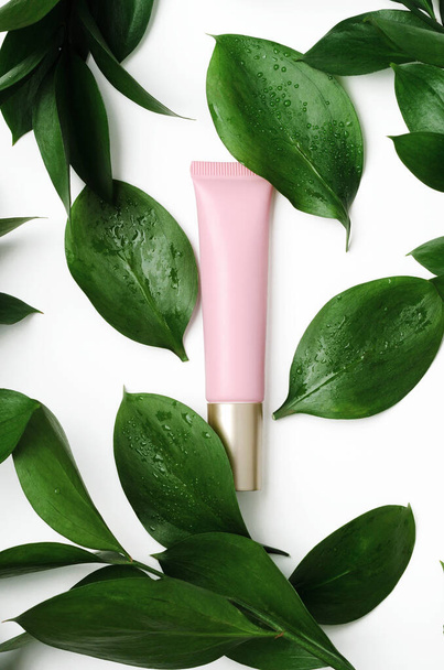 Cosmetology tube with green leaves on white background. Top class facial care serum with decorative fresh ficus foliage and dew drops botanical backdrop. Eye cream, concealer in pink tube promo. - Photo, Image