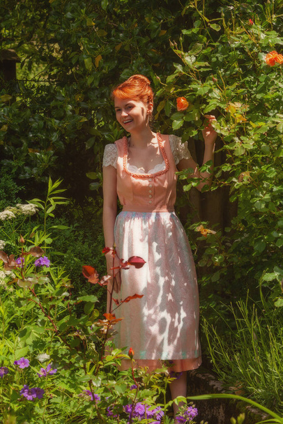 Nuori nainen dirndl nauraa puutarhassa ja iloisesti pitäen ruusu kädessään. - Valokuva, kuva