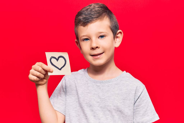 Cute blond kid holding heart reminder looking positive and happy standing and smiling with a confident smile showing teeth  - Foto, imagen
