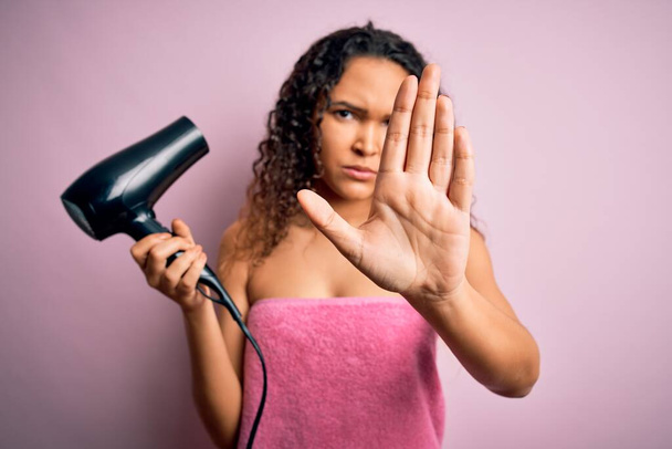 Красивая женщина с вьющимися волосами носить душ полотенце после ванны с помощью фена с открытой рукой делает знак стоп с серьезным и уверенным выражением, защитный жест - Фото, изображение