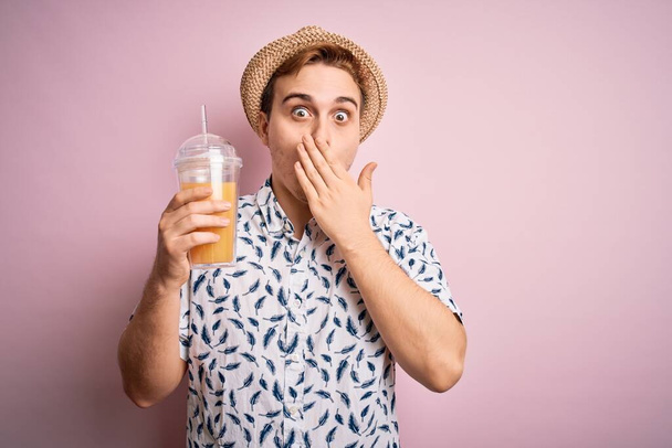 Nuori komea punapää mies lomalla juomassa lasia terveellistä appelsiinimehua, joka peittää suun käsin, järkyttynyt ja pelkää erehdystä. Yllättynyt ilme - Valokuva, kuva