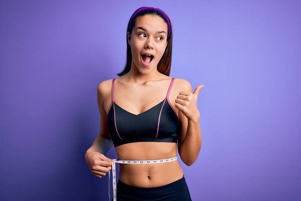 Beautiful slim girl controlling weight using measure tape on waist over purple background pointing and showing with thumb up to the side with happy face smiling - 写真・画像