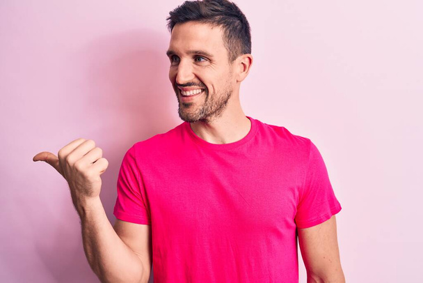Młody przystojny mężczyzna ubrany w luźny t-shirt stojący nad odizolowanym różowym tłem wskazując kciuk do góry na bok uśmiechnięty szczęśliwy z otwartymi ustami - Zdjęcie, obraz