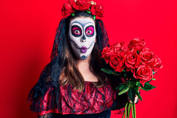 Jeune femme portant le jour du costume mort tenant des fleurs effrayées et étonnées avec la bouche ouverte pour la surprise, visage incrédule  - Photo, image