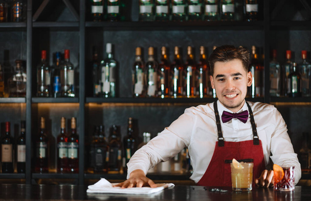 Портрет дружелюбного улыбающегося молодого бармена, стоящего у барной стойки - Фото, изображение