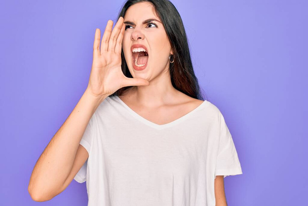 Young beautiful brunette woman wearing casual white t-shirt over purple background shouting and screaming loud to side with hand on mouth. Communication concept. - Фото, зображення