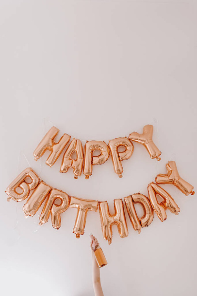 Female hand hold bottle of rose champagne. Rose gold balloons Happy Birthday on white wall. Anniversary party celebrating decoration - Photo, Image