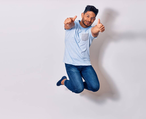 Young handsome latin man wearing casual clothes smiling happy. Jumping with smile on face doing ok sign with thumbs up over isolated white background - Fotó, kép