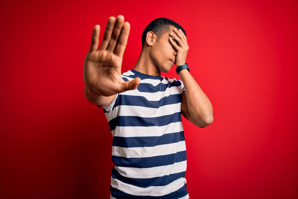Schöner afrikanisch-amerikanischer Mann in lässig gestreiftem T-Shirt, der über rotem Hintergrund steht und die Augen mit Händen bedeckt und mit traurigem und ängstlichem Gesichtsausdruck Stopp-Gesten macht. Peinliches und negatives Konzept. - Foto, Bild