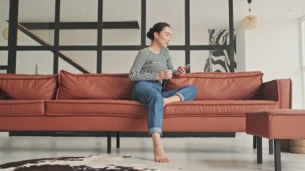 Smiling pensive pretty asian woman using smartphone then looking away while sitting on sofa at home - Filmmaterial, Video
