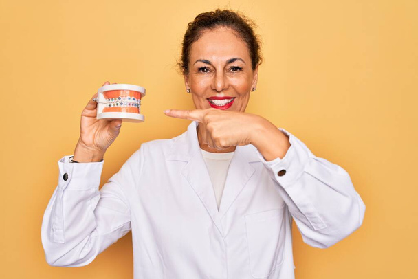 Média idade dentista sênior mulher segurando ortodontia dentadura com aparelho aligner muito feliz apontando com mão e dedo
 - Foto, Imagem