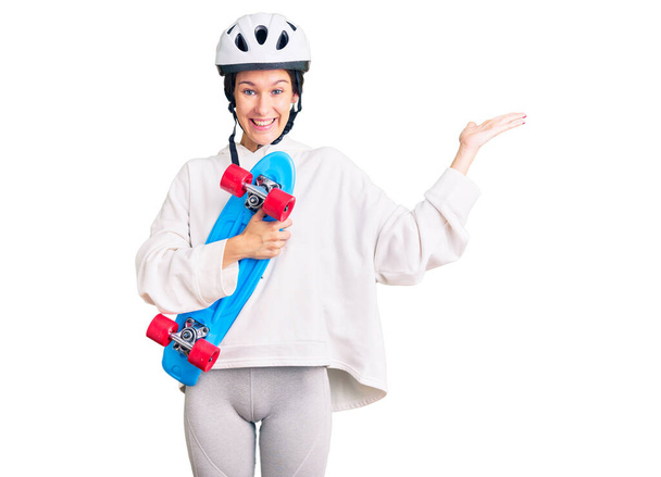Mooie brunette jonge vrouw dragen veiligheid helm en schaatsen vieren overwinning met een vrolijke glimlach en winnaar uitdrukking met opgeheven handen  - Foto, afbeelding