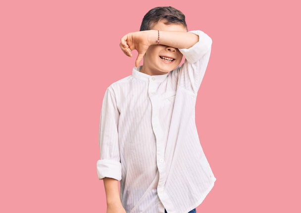 Cute blond kid wearing elegant shirt covering eyes with arm smiling cheerful and funny. blind concept.  - Foto, imagen