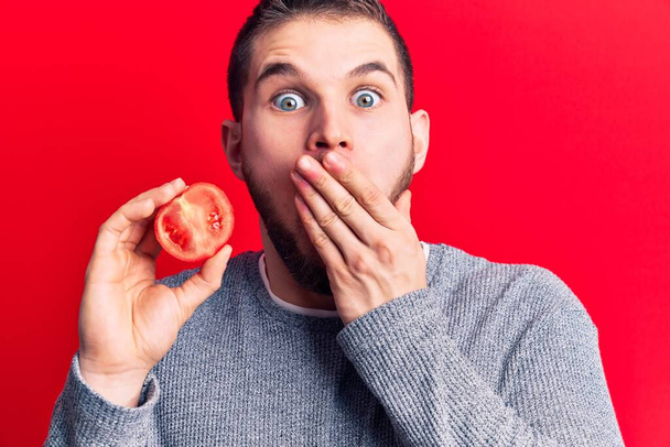 Young handsome man holding slice of tomato covering mouth with hand, shocked and afraid for mistake. surprised expression  - Photo, Image