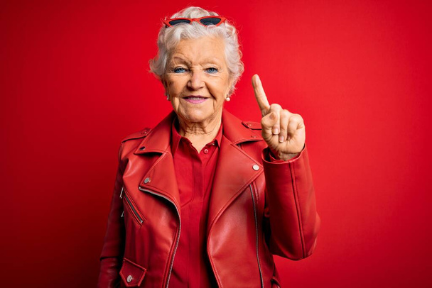 Senior kaunis harmaa tukka nainen yllään rento punainen takki ja aurinkolasit näy ja osoittaa sormella numero yksi hymyillen itsevarma ja onnellinen. - Valokuva, kuva