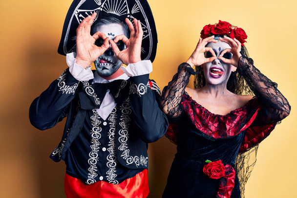 Young couple wearing mexican day of the dead costume over yellow doing ok gesture like binoculars sticking tongue out, eyes looking through fingers. crazy expression.  - Photo, Image