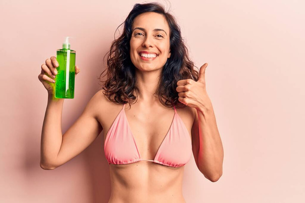 Jonge mooie Latijns-Amerikaanse vrouw draagt bikini met aloë vera lotion glimlachen gelukkig en positief, duim omhoog doen uitstekend en goedkeuring teken  - Foto, afbeelding