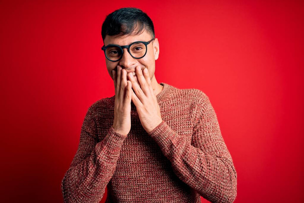 Jovem hispânico bonito vestindo óculos nerd sobre fundo vermelho rindo e riso embaraçado cobrindo a boca com as mãos, fofocas e conceito escândalo
 - Foto, Imagem