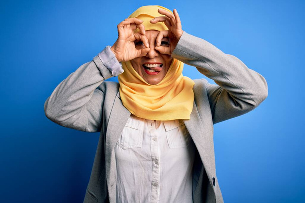 Donna d'affari bruna di mezza età che indossa hijab tradizionale musulmano su sfondo blu facendo un gesto ok come un binocolo che sporge la lingua, gli occhi che guardano attraverso le dita. Espressione folle. - Foto, immagini