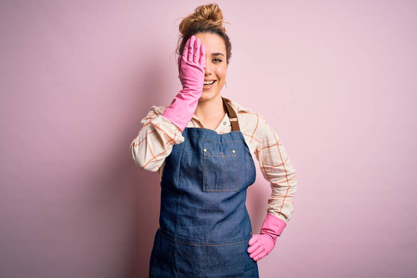 Mladá krásná blondýnka uklízečka dělá domácí práce nosit arpon a rukavice zakrývající jedno oko s rukou, sebevědomý úsměv na tváři a překvapivé emoce. - Fotografie, Obrázek