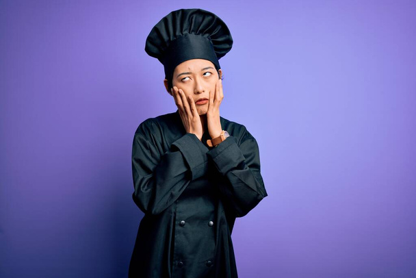 若い美しい中国のシェフの女性は、調理器の制服と紫色の背景の上に帽子をかぶって顔、うつ病や悲しみ、動揺や問題のためにイライラをカバー疲れた手 - 写真・画像