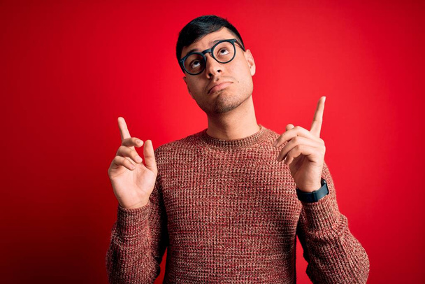Jovem hispânico bonito vestindo óculos nerd sobre fundo vermelho Apontando para cima olhar triste e chateado, indicando direção com os dedos, infeliz e deprimido
. - Foto, Imagem