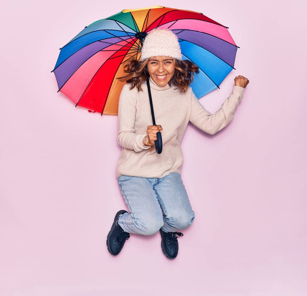 Young beautiful latin woman holding umbrella smiling happy. Jumping with smile on face over isolated pink background - 写真・画像
